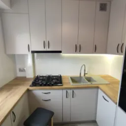 Jasna kuchnia z drewnianym blatem, czarnymi uchwytami i szarym zlewem. Na blacie kuchenka gazowa. Pod blatem szary taboret.