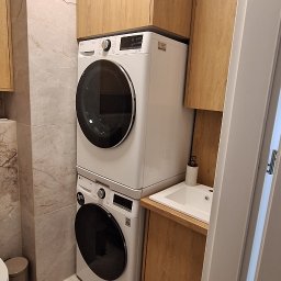 Piotr Skóra - Jasna łazienka z zabudową meblową z drewnianymi lamelami na suficie. Pralka i suszarka w słupku obok umywalki z szafką. Marmurowe płytki na ścianach i podłodze.