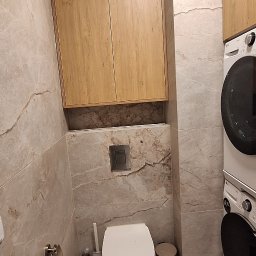 Piotr Skóra - Nowoczesna toaleta z szafką wiszącą z drewna i pralką w tle. Beżowe płytki imitujące marmur na ścianach i podłodze. Minimalistyczny design.