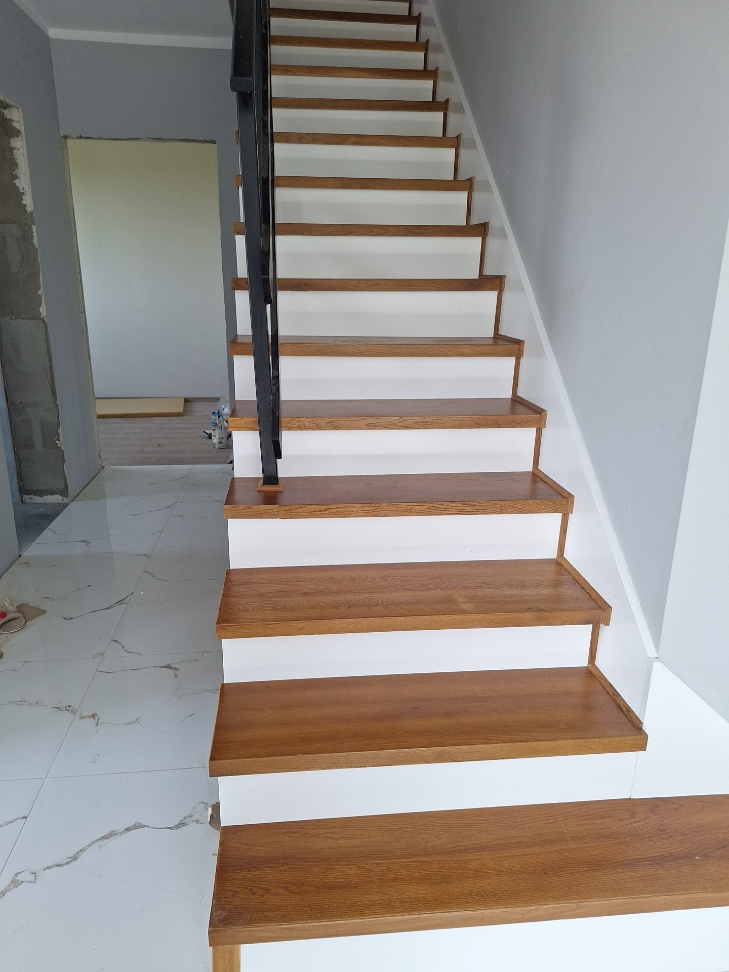 Nowoczesne schody drewniane z białymi podstopnicami i czarną balustradą, widok z dołu. Wykończenie wnętrza w trakcie realizacji, marmurowa podłoga.