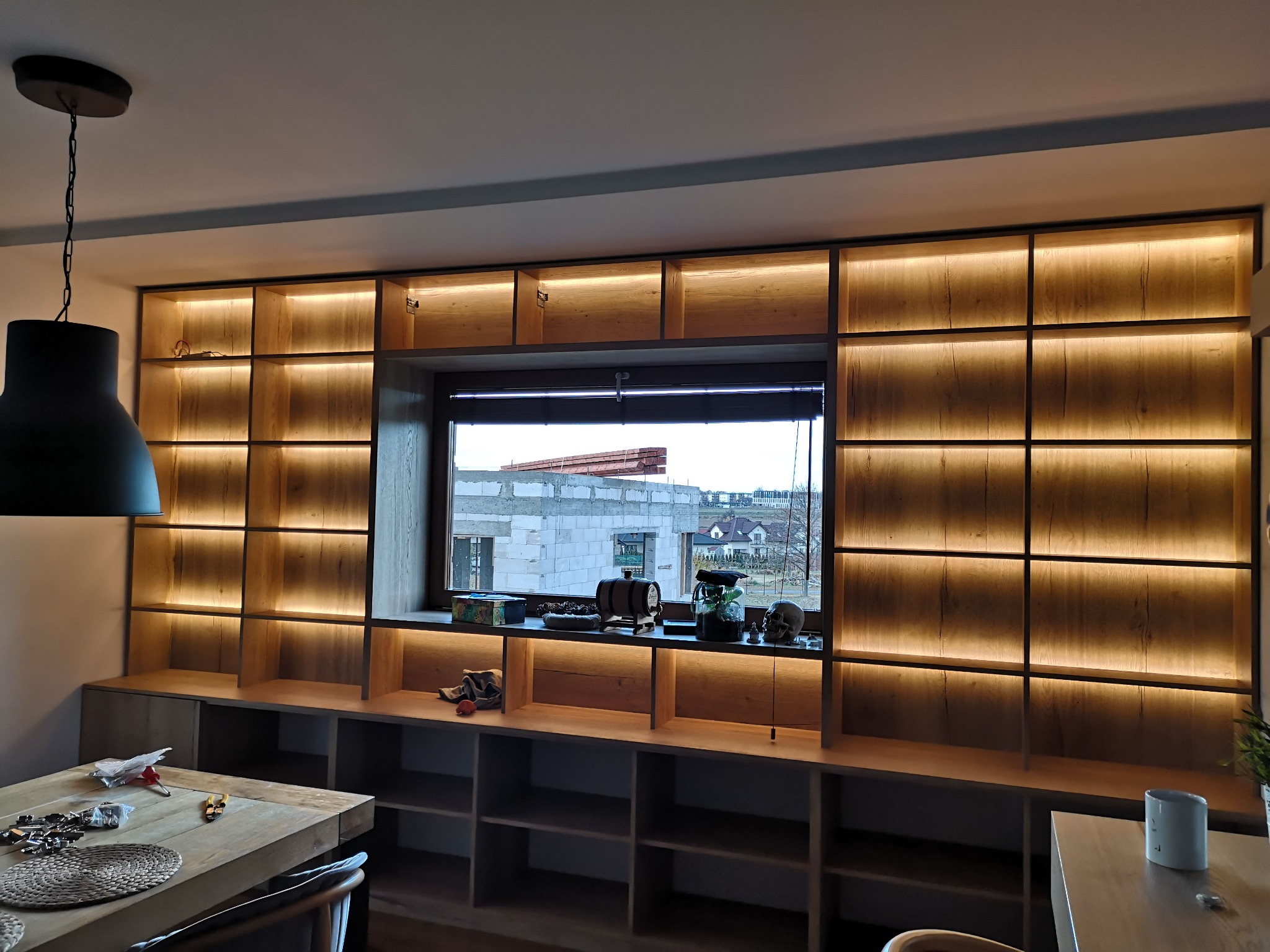 Drewniana zabudowa meblowa z oświetleniem LED, zintegrowana z oknem, widok na budowę za oknem. Na pierwszym planie stół z elementami montażowymi.