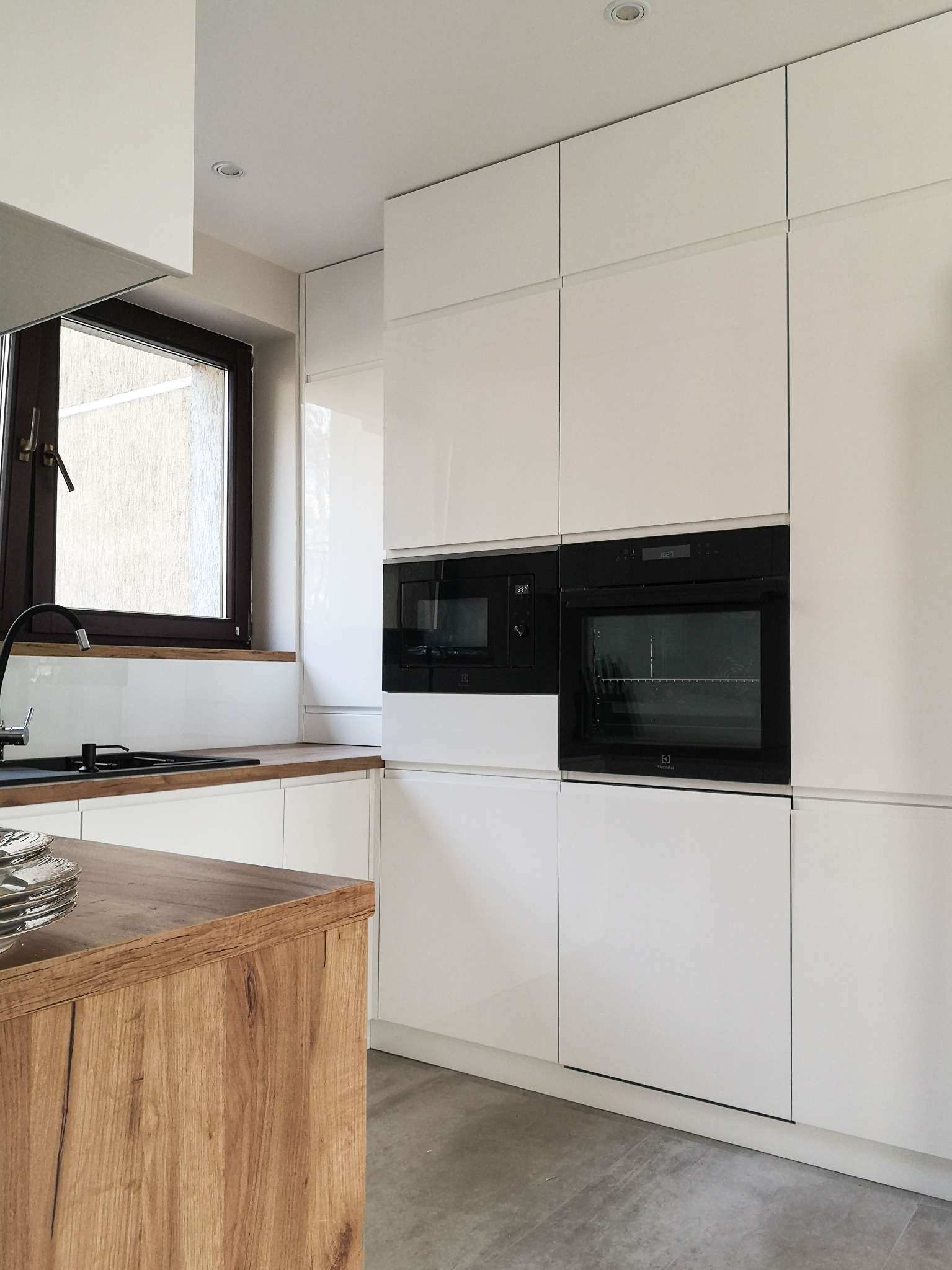 Nowoczesna kuchnia z białymi, lakierowanymi frontami, drewnianym blatem i czarnymi urządzeniami AGD w zabudowie. Minimalistyczny design, szare płytki podłogowe.