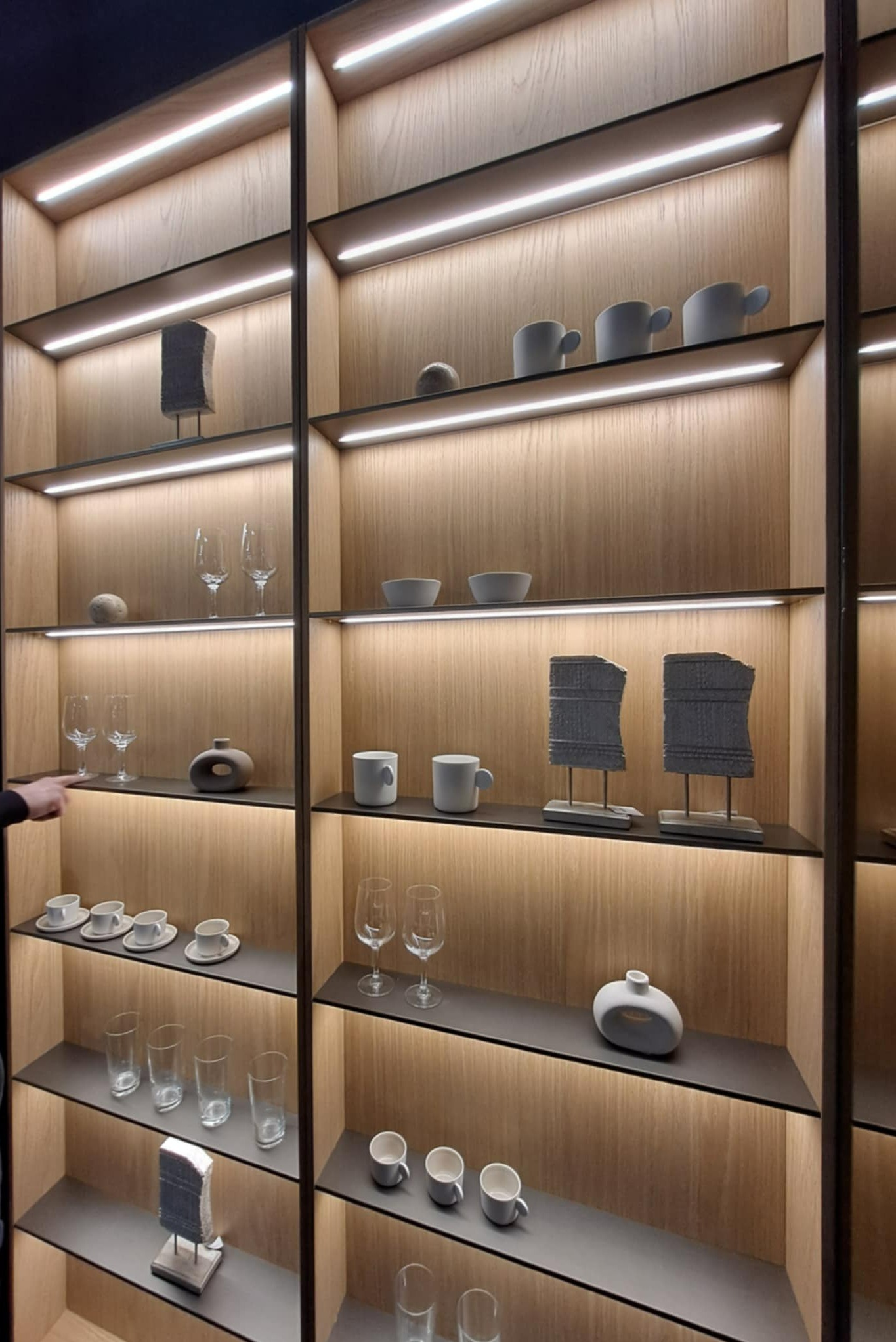 Podświetlana, zabudowana półka z drewnianym wykończeniem, na której znajdują się dekoracyjne przedmioty, szklanki i filiżanki. Nowoczesny design z subtelnym oświetleniem LED.