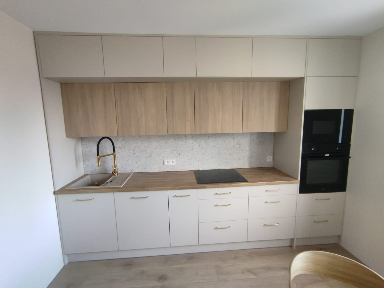 Jasna kuchnia z drewnianymi akcentami: białe szafki, blat z drewna, złota bateria i zabudowany piekarnik. Minimalistyczny design z terazzo na ścianie.