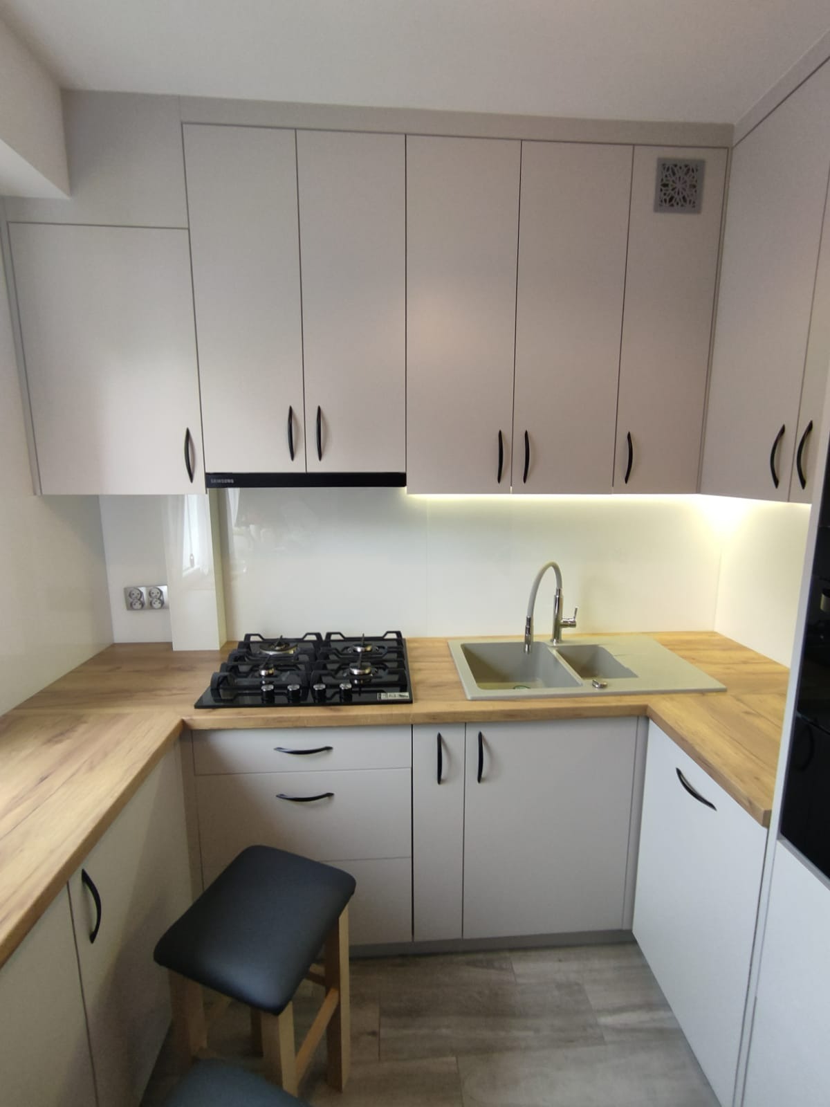 Jasna kuchnia z drewnianym blatem, czarnymi uchwytami i szarym zlewem. Na blacie kuchenka gazowa. Pod blatem szary taboret.