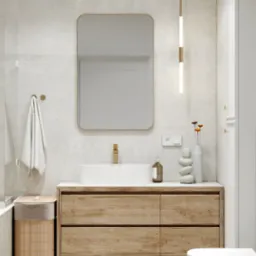 Jasna łazienka z drewnianą szafką pod umywalką, minimalistycznym lustrem w złotej ramie i wiszącą lampą. Umywalka nablatowa, dekoracje i kosz z bambusa.