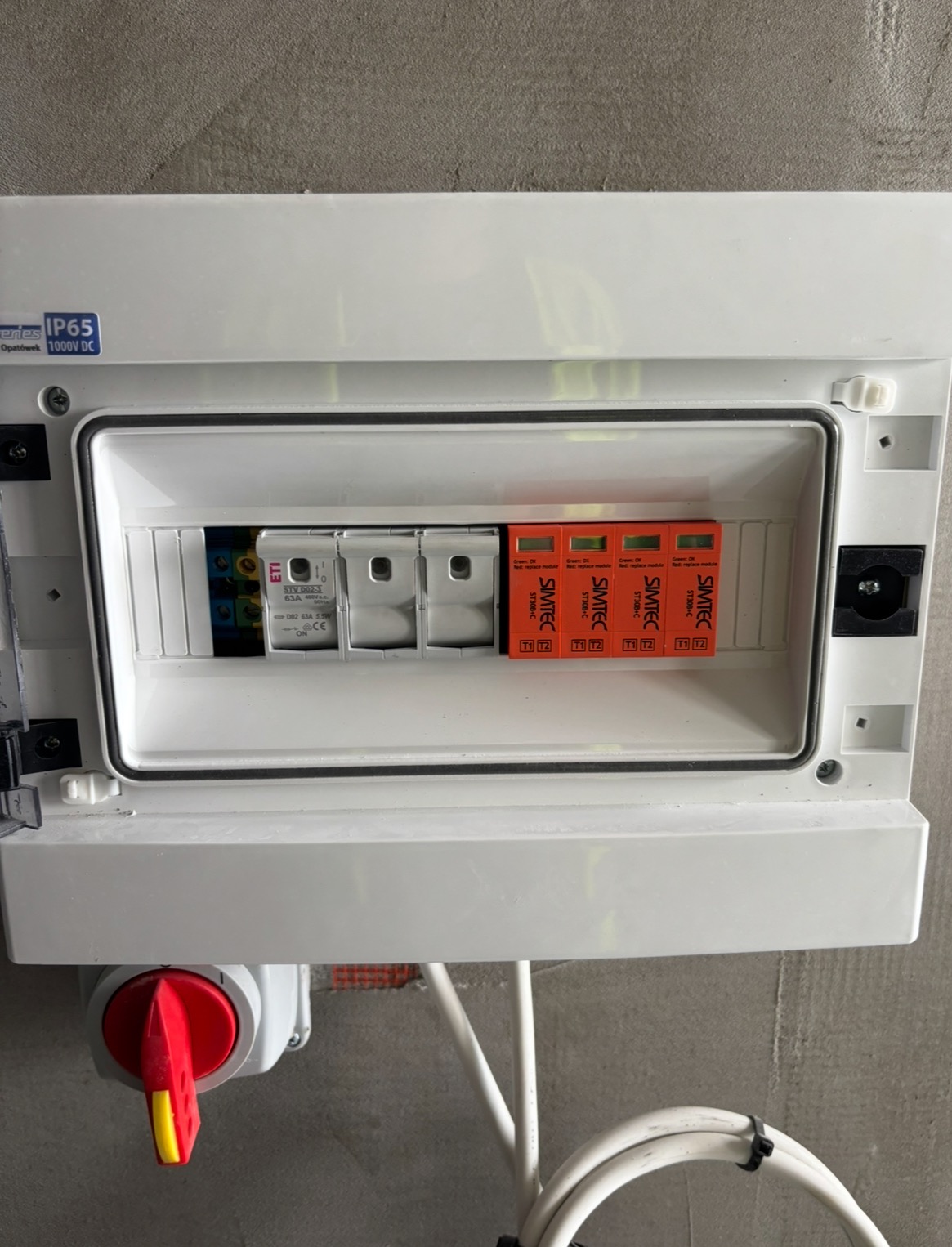 Otwarta, biała skrzynka elektryczna z widocznymi bezpiecznikami i wyłącznikiem. Widoczne kable. Skrzynka zamontowana na szarej ścianie.