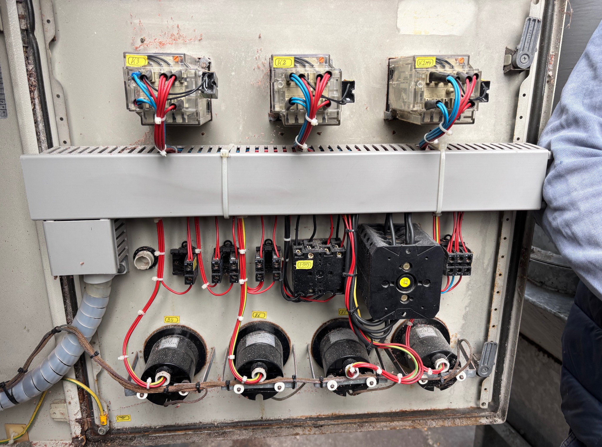 Otwarta skrzynka elektryczna z widocznymi przekaźnikami, przewodami w kolorach czerwonym, niebieskim i czarnym. Elementy oznaczone żółtymi etykietami. Widoczne ślady użytkowania.