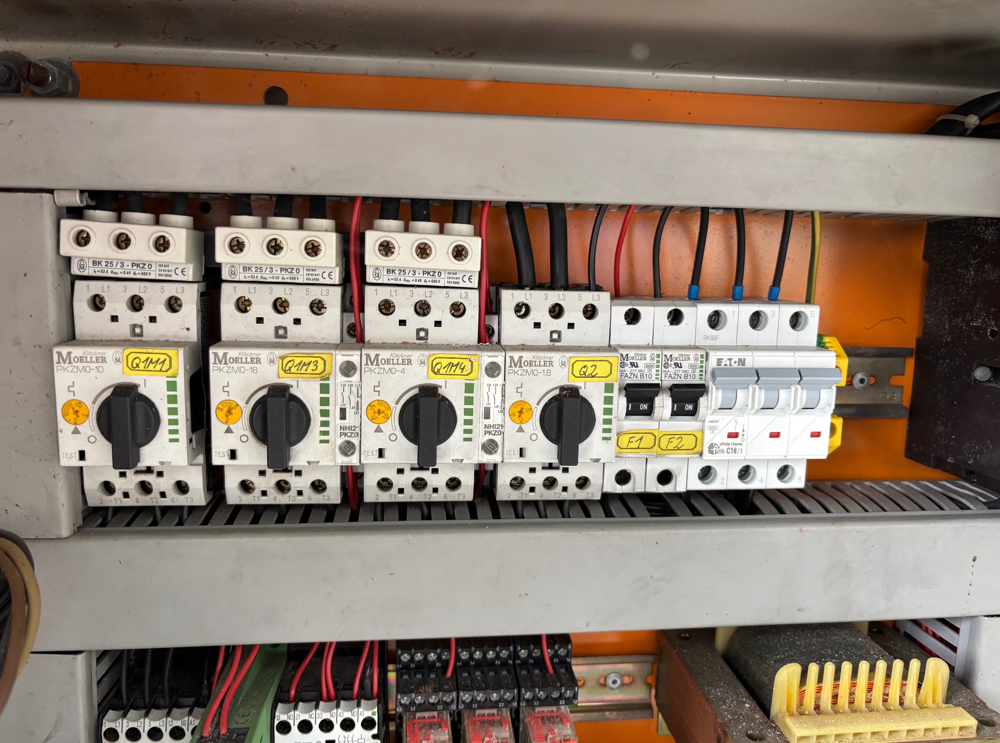 Rozdzielnica elektryczna z bezpiecznikami, wyłącznikami i przewodami. Widoczne elementy sterujące i oznaczenia. Pomarańczowe tło wewnątrz szafy.