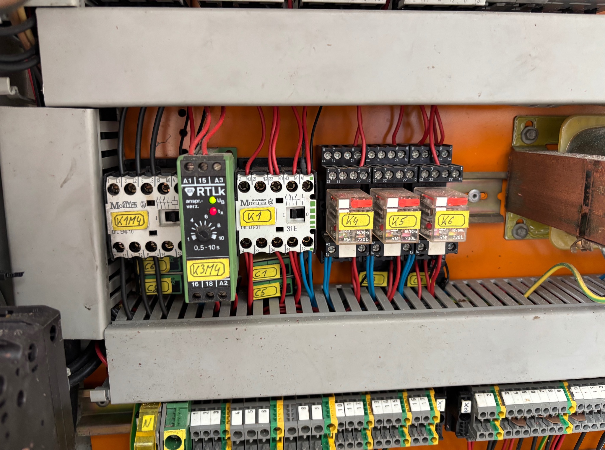 Rozdzielnica elektryczna z widocznymi elementami sterującymi, przekaźnikami i okablowaniem. Elementy oznaczone żółtymi etykietami. Widoczne złącza i przewody.