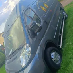 Szary bus dostawczy marki Peugeot z logo firmy transportowej 'Partykar Verhuur' na tle zielonej trawy i błękitnego nieba z chmurami. Ujęcie z dołu.