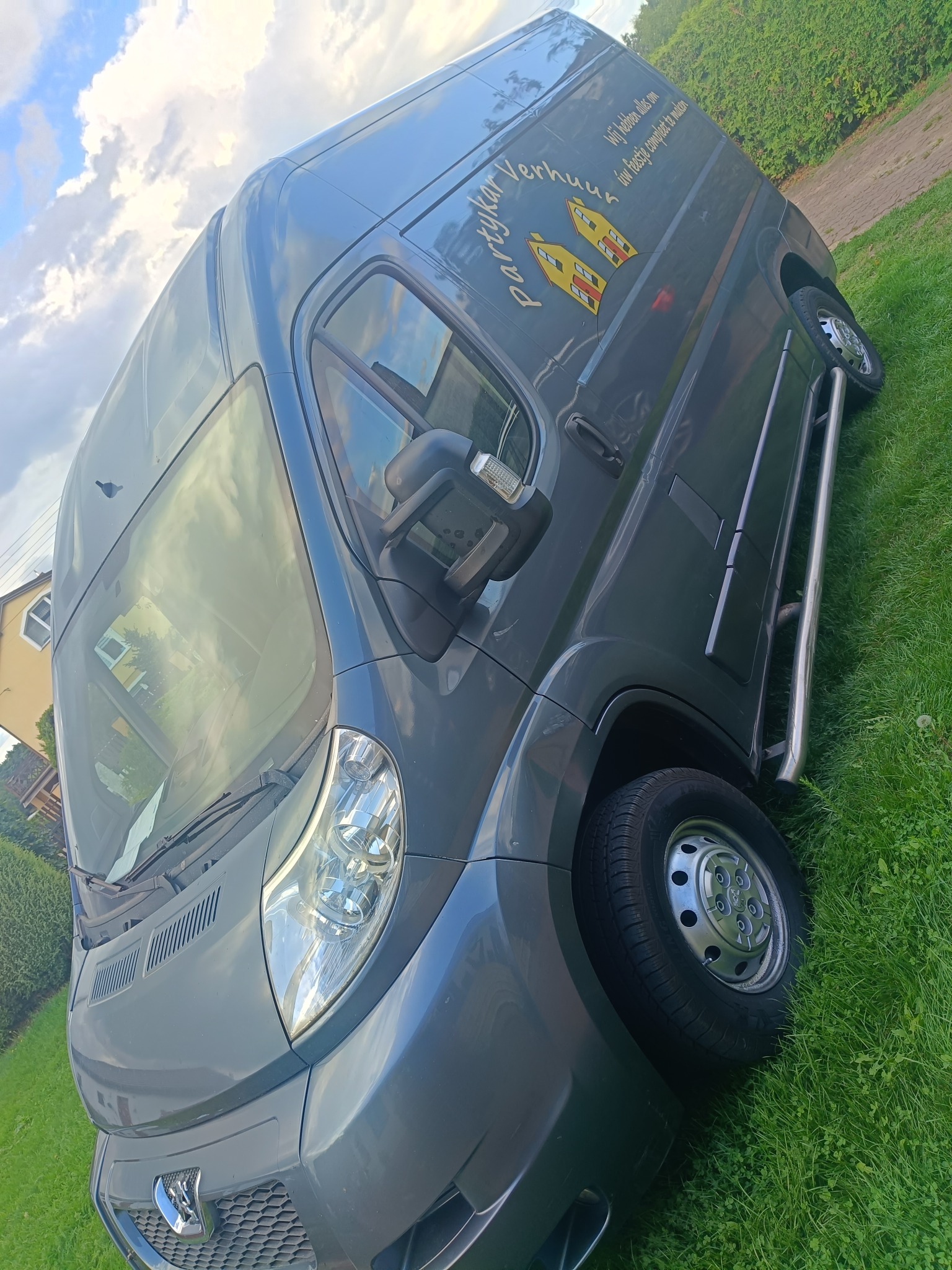 Szary bus dostawczy marki Peugeot z logo firmy transportowej 'Partykar Verhuur' na tle zielonej trawy i błękitnego nieba z chmurami. Ujęcie z dołu.