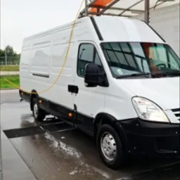 Biały bus Iveco w myjni automatycznej w Wejherowie, widok z boku, w trakcie mycia. Woda i piana na posadzce. Żółty wąż myjący zwisa z góry.