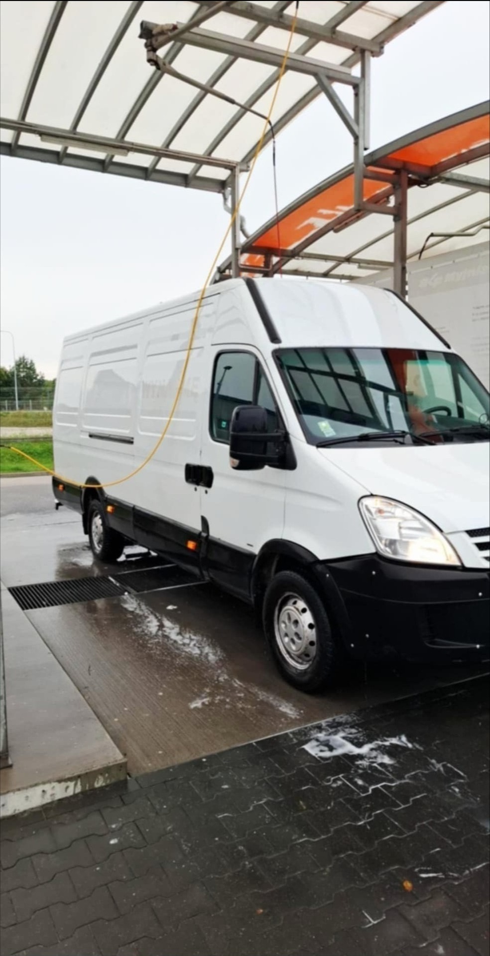 Biały bus Iveco w myjni automatycznej w Wejherowie, widok z boku, w trakcie mycia. Woda i piana na posadzce. Żółty wąż myjący zwisa z góry.