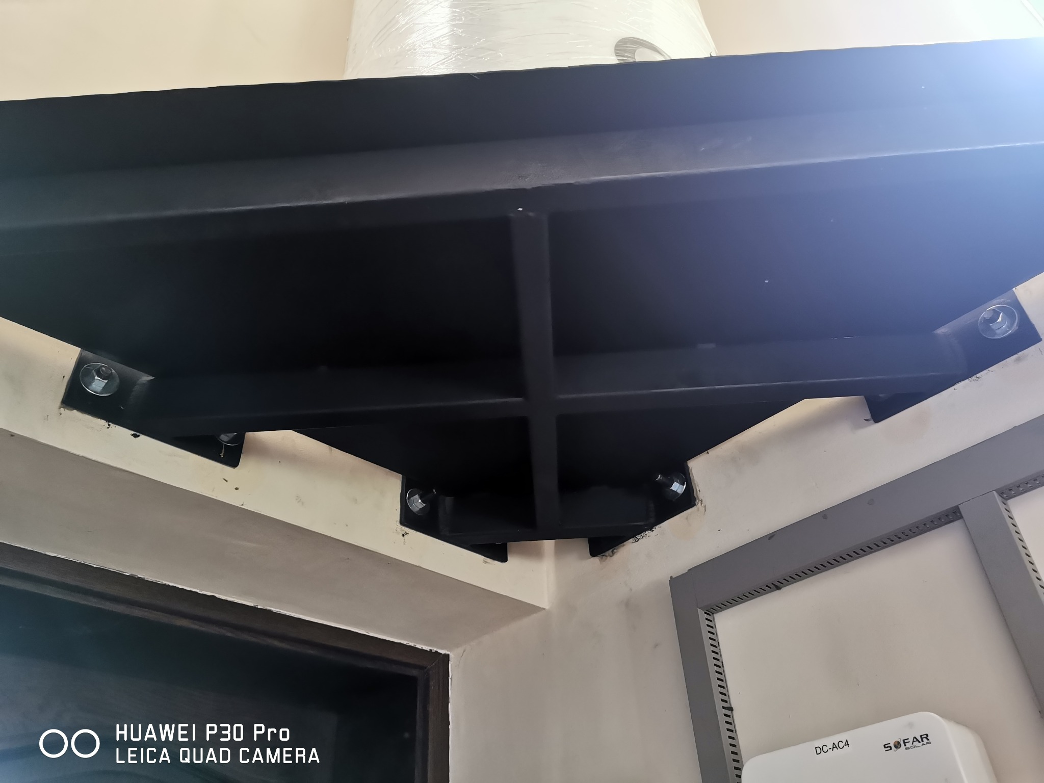 Czarna, metalowa konstrukcja wspornikowa przymocowana do ściany za pomocą śrub, widoczna z dołu. Element instalacji wewnątrz budynku, blisko drzwi i szafki.