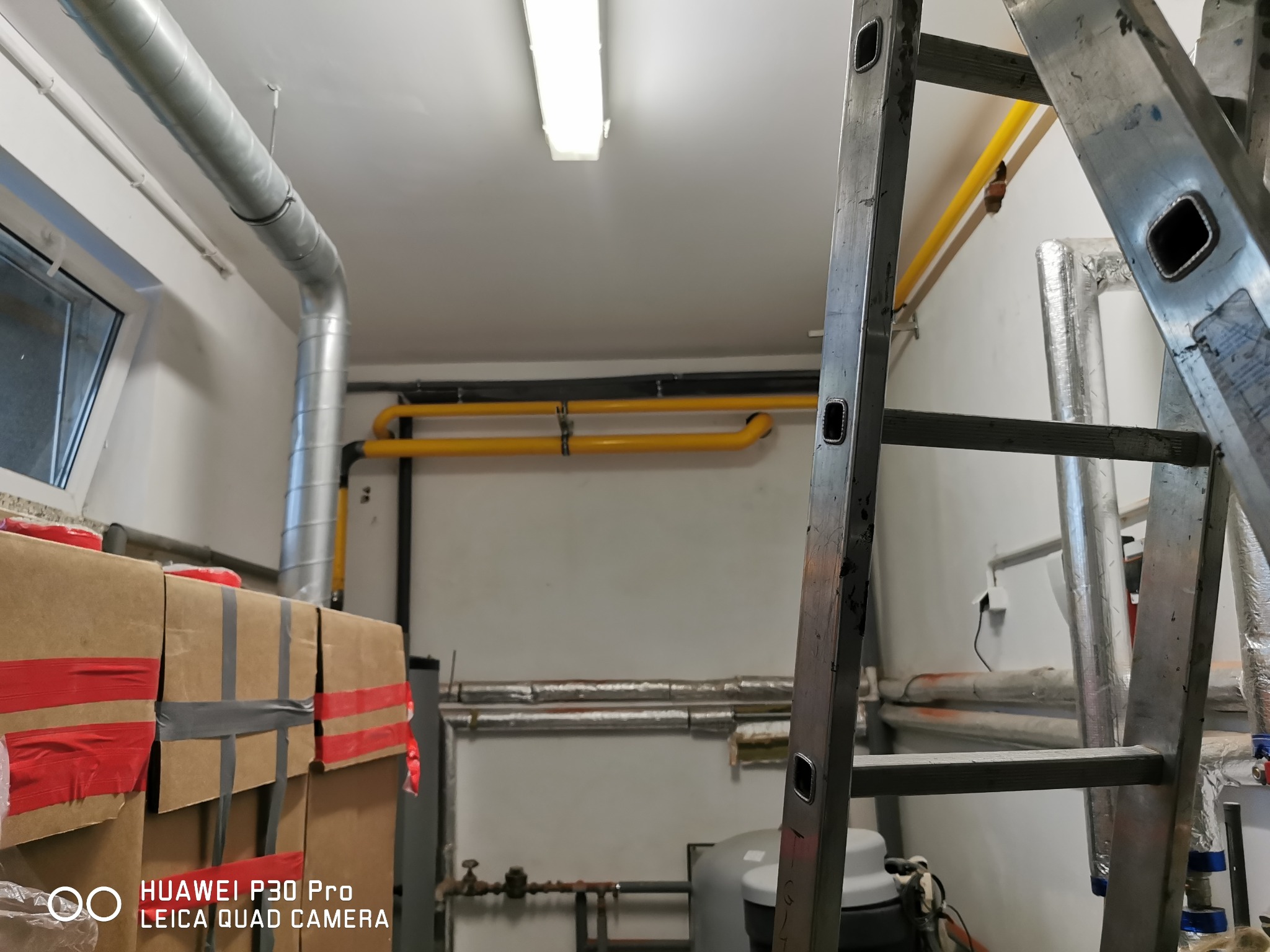 Wnętrze kotłowni z żółtymi rurami gazowymi, srebrnymi rurami wentylacyjnymi i grzewczymi, oraz aluminiową drabiną. Widoczne kartony z materiałami.