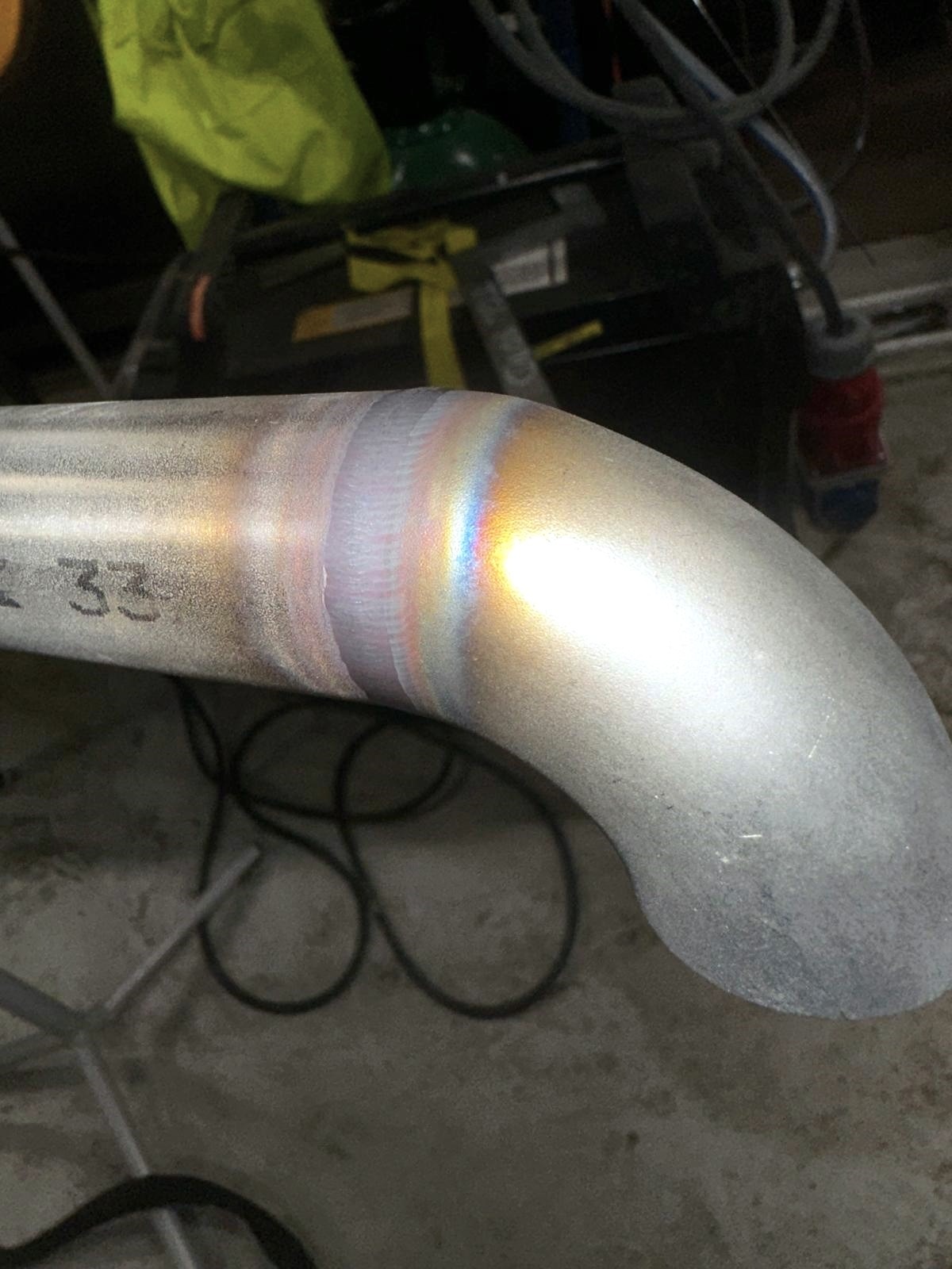 Metalowa rura po spawaniu z widocznymi kolorami spoiny, leżąca na warsztatowym stole. Widoczne ślady obróbki i numer '2 33' na powierzchni.