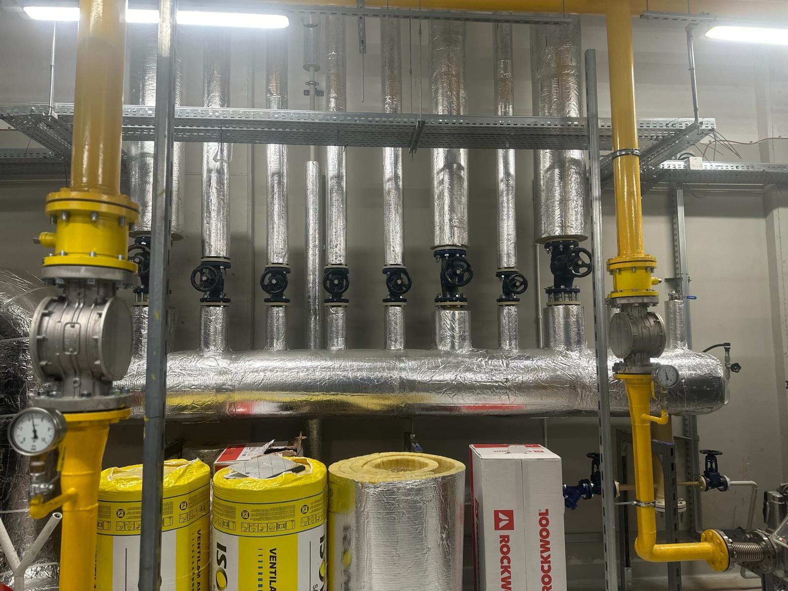 Izolowane rury gazowe z zaworami i manometrami, żółte rury pionowe, materiały izolacyjne Rockwool i ISO Ventil w tle, szara ściana, oświetlenie sufitowe.