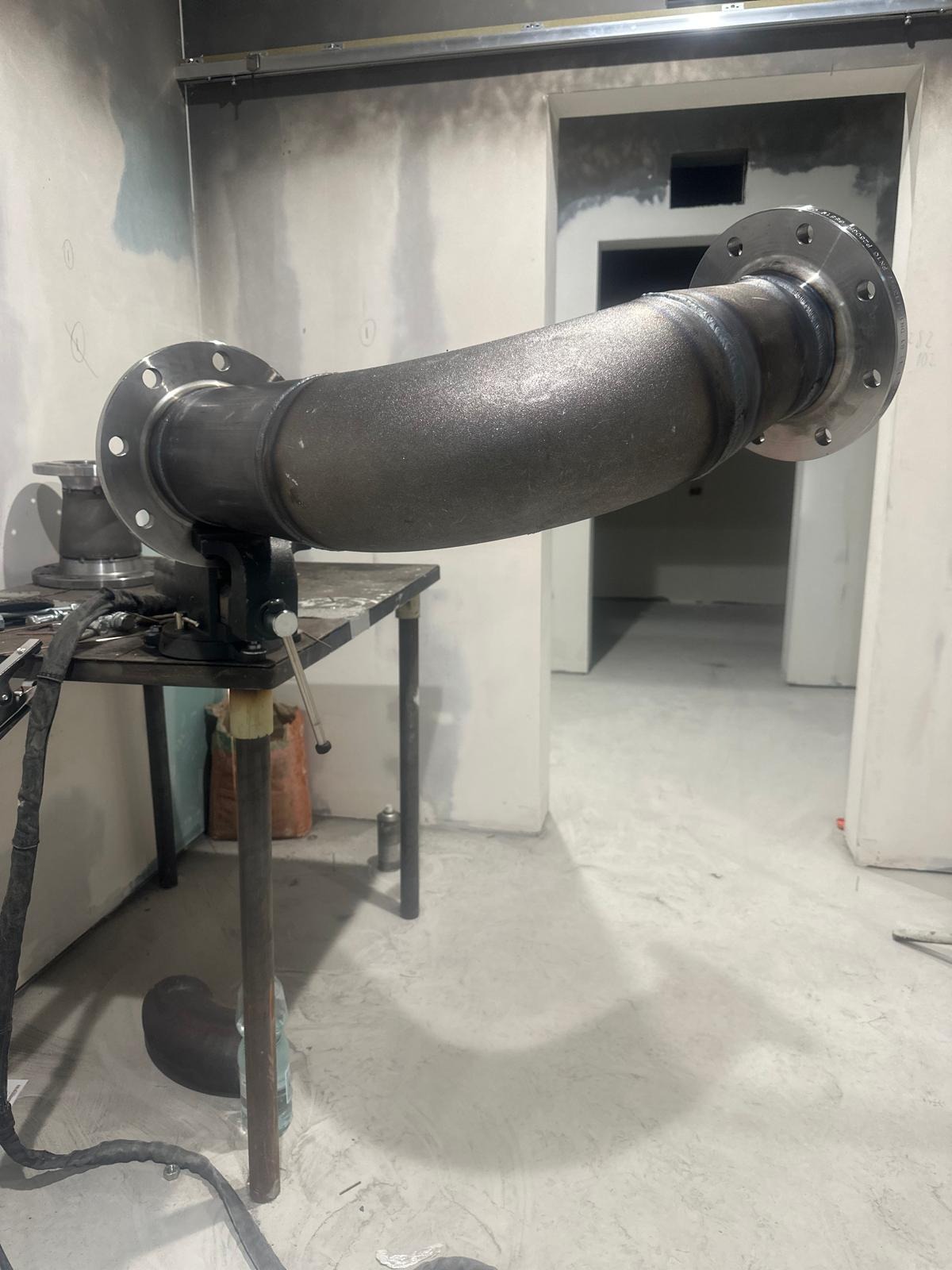 Spawany, zakrzywiony element rury z kołnierzami, zamocowany w imadle na stole warsztatowym. Widoczne ślady spawania i surowe wykończenie. Tło to surowe ściany w trakcie remontu.