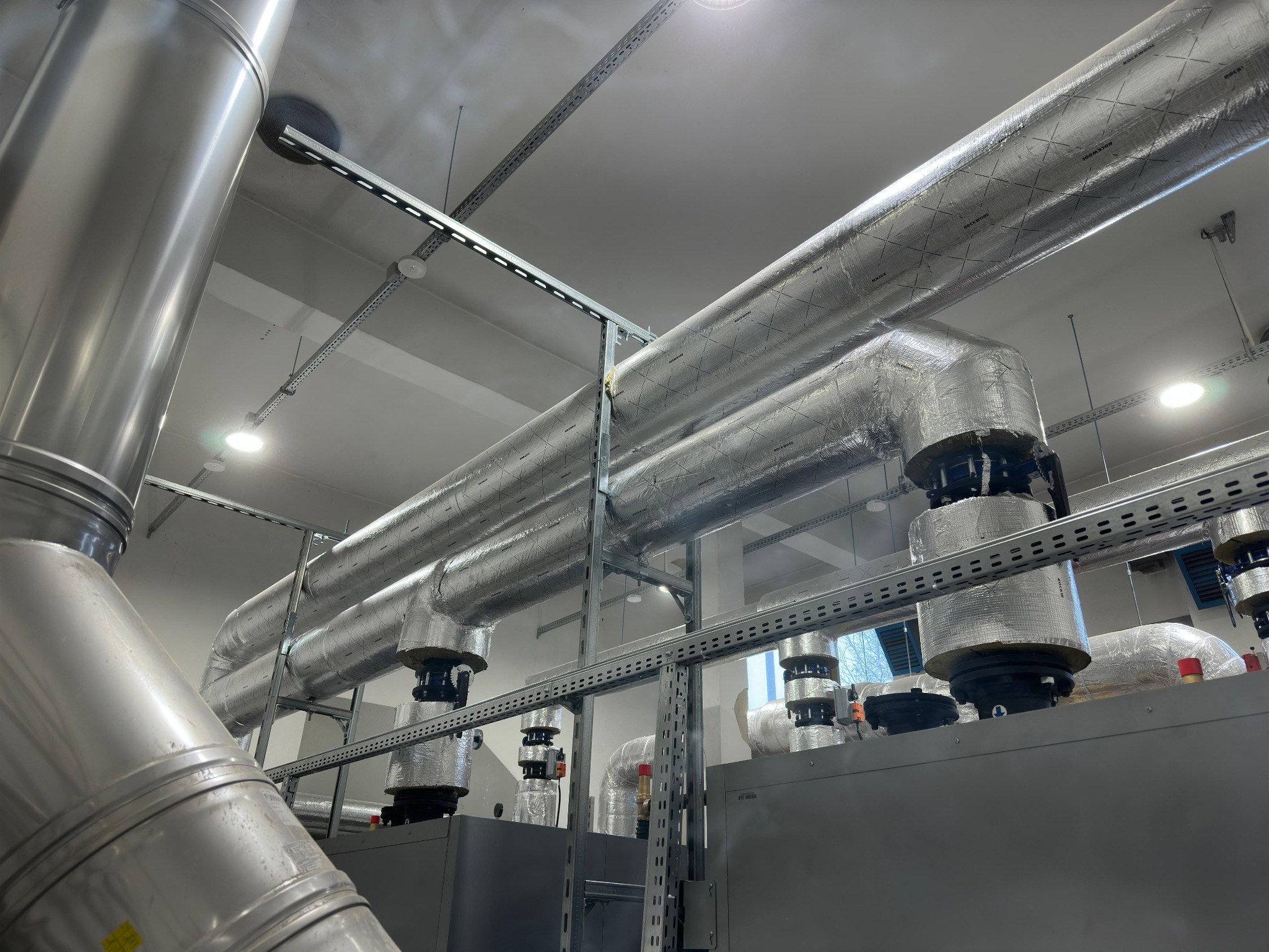 System rur wentylacyjnych z izolacją, wsparty na metalowej konstrukcji, w jasnym pomieszczeniu. Widoczne elementy mocujące i oświetlenie sufitowe.