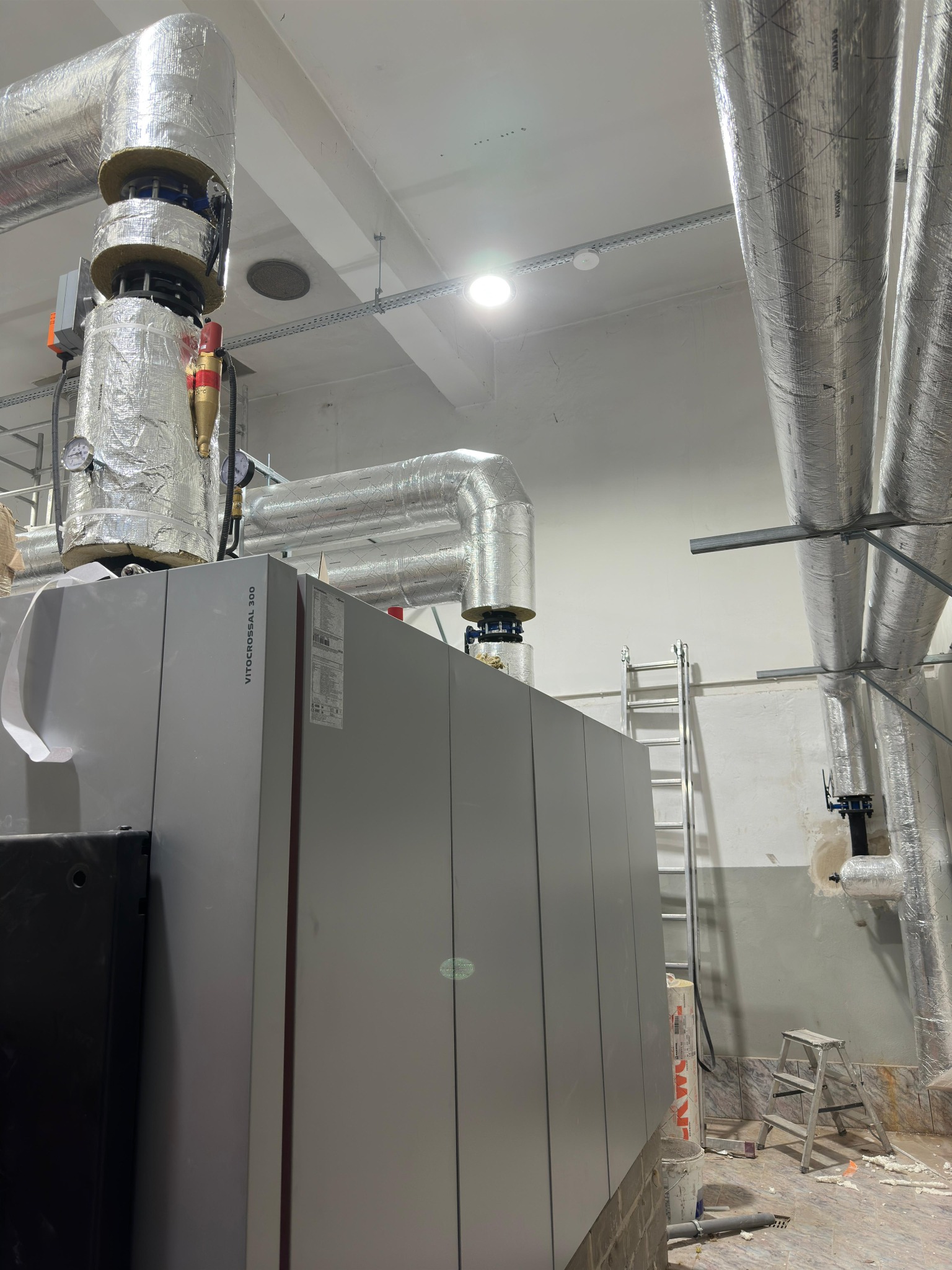 Szary piec Viessmann Vitocrossal 300 z izolowanymi rurami w pomieszczeniu technicznym, drabina i schodki w tle. Widoczne elementy instalacji.