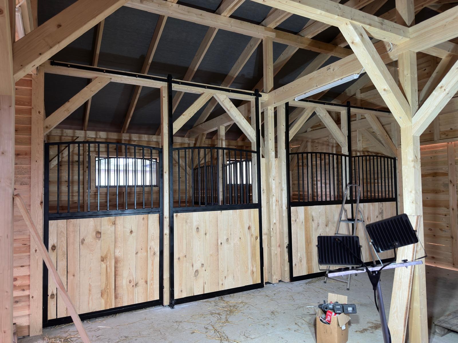 Wnętrze stajni w trakcie budowy z drewnianą konstrukcją i metalowymi przegrodami dla koni. Widoczne narzędzia i oświetlenie budowlane.