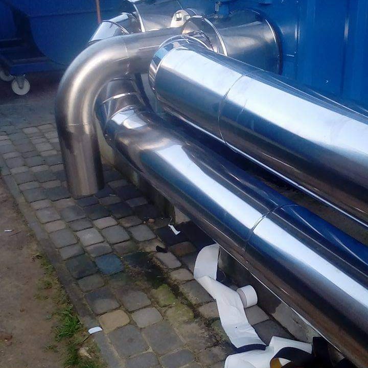 Błyszczące, metalowe rury instalacji przemysłowej ułożone na brukowanej powierzchni, z widocznymi elementami izolacji i fragmentem niebieskiej konstrukcji w tle.