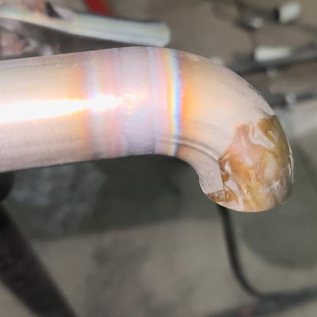 Błyszczący, zakrzywiony fragment metalowej rury, prawdopodobnie ze stali nierdzewnej, z widocznymi śladami obróbki i tęczowymi refleksami świetlnymi.