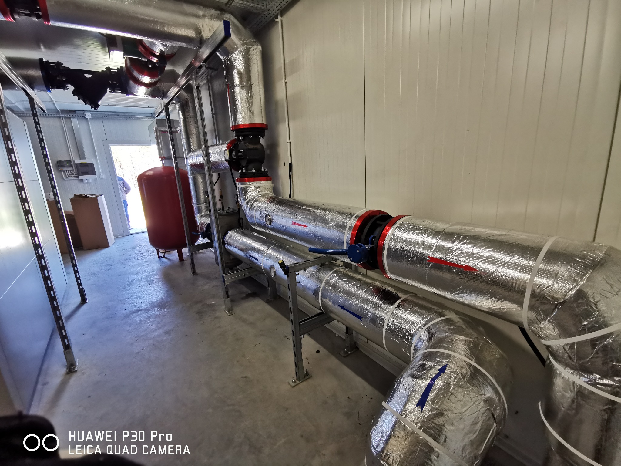 Izolowane rury instalacji wewnątrz pomieszczenia z widocznym zbiornikiem wyrównawczym i fragmentem osoby w tle, ukazujące etap prac instalacyjnych.