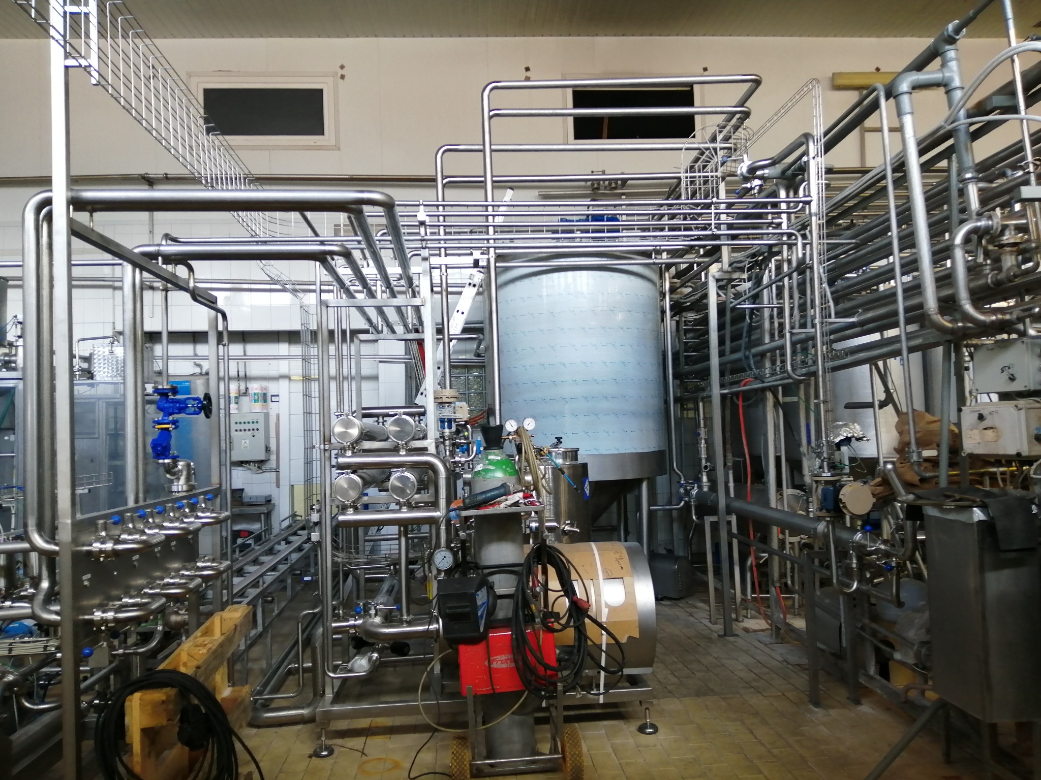 Kompleksowa instalacja przemysłowa z rurami ze stali nierdzewnej, zbiornikiem i aparaturą kontrolną w jasnym pomieszczeniu. Widoczne elementy spawarki na wózku.