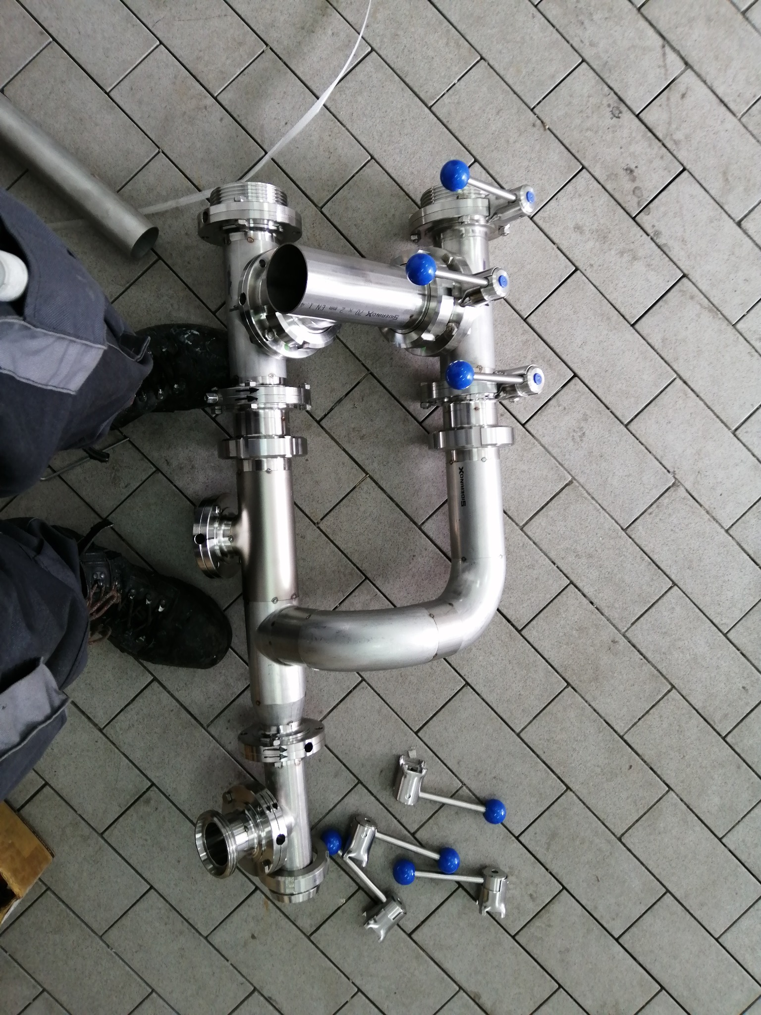 Elementy instalacji ze stali nierdzewnej z niebieskimi zaworami na szarych płytkach, widoczne fragmenty odzieży roboczej i rury. Nietypowe ułożenie instalacji.