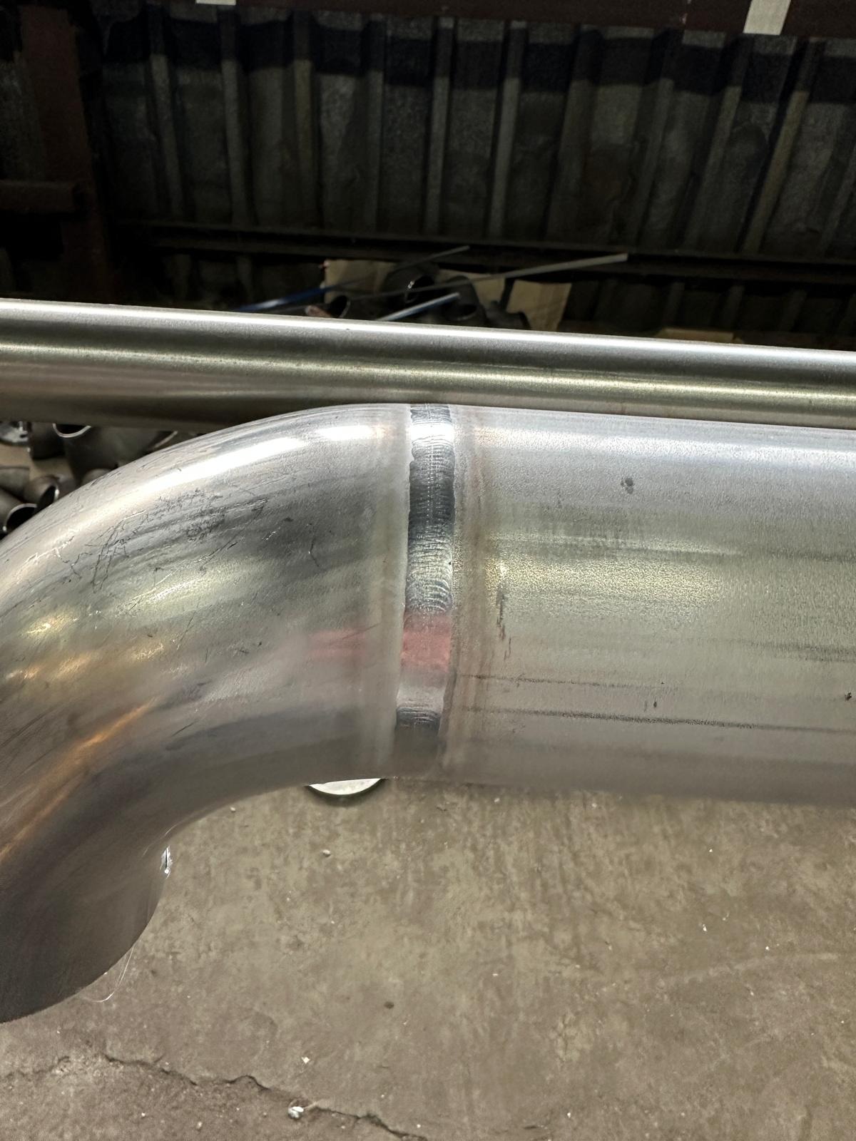 Szlifowany, spawany element rury ze stali nierdzewnej, widoczny spaw na zagięciu. Metaliczna powierzchnia z refleksami światła, surowe tło warsztatowe.