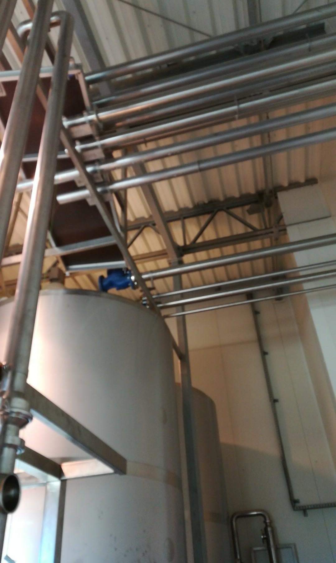 Metalowe rury i zbiorniki w hali produkcyjnej, widok z dołu. Elementy konstrukcyjne pod sufitem. Niebieski zawór na jednym ze zbiorników.