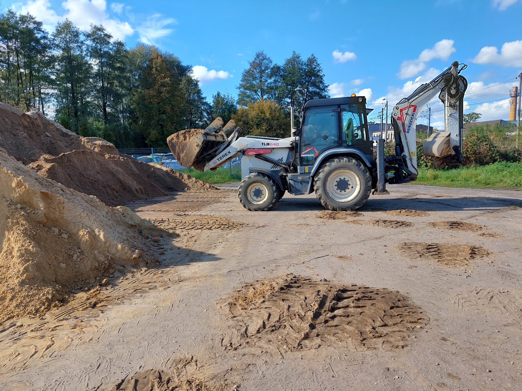 Biała koparko-ładowarka Terex TLB840 z pełną łyżką piasku na placu budowy, ślady opon na ziemi, w tle drzewa i błękitne niebo.