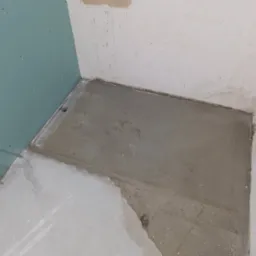 Przygotowana podłoga w łazience z odpływem liniowym, ściany w stanie surowym, płyta GK. Widoczny etap prac przed położeniem płytek. Szary beton i surowe wykończenie.