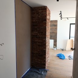 Paulina Kubicka Mateusz Banaszak - Wykończony filar z cegły klinkierowej w nowym mieszkaniu, zabezpieczony folią malarską. Ściany przygotowane do malowania, widoczne taśmy malarskie.
