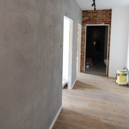 Paulina Kubicka Mateusz Banaszak - Szara ściana z efektem dekoracyjnym, drewnopodobne panele podłogowe, fragment ściany z cegły i odkurzacz w tle. Widok na wykończone wnętrze.
