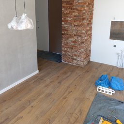 Paulina Kubicka Mateusz Banaszak - Podłoga z paneli w trakcie układania, widoczne narzędzia, folia ochronna, fragment kolumny z cegły i osłonięte lampy. Etap prac wykończeniowych.