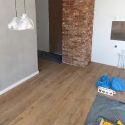 Podłoga z paneli w trakcie układania, widoczne narzędzia, folia ochronna, fragment kolumny z cegły i osłonięte lampy. Etap prac wykończeniowych.