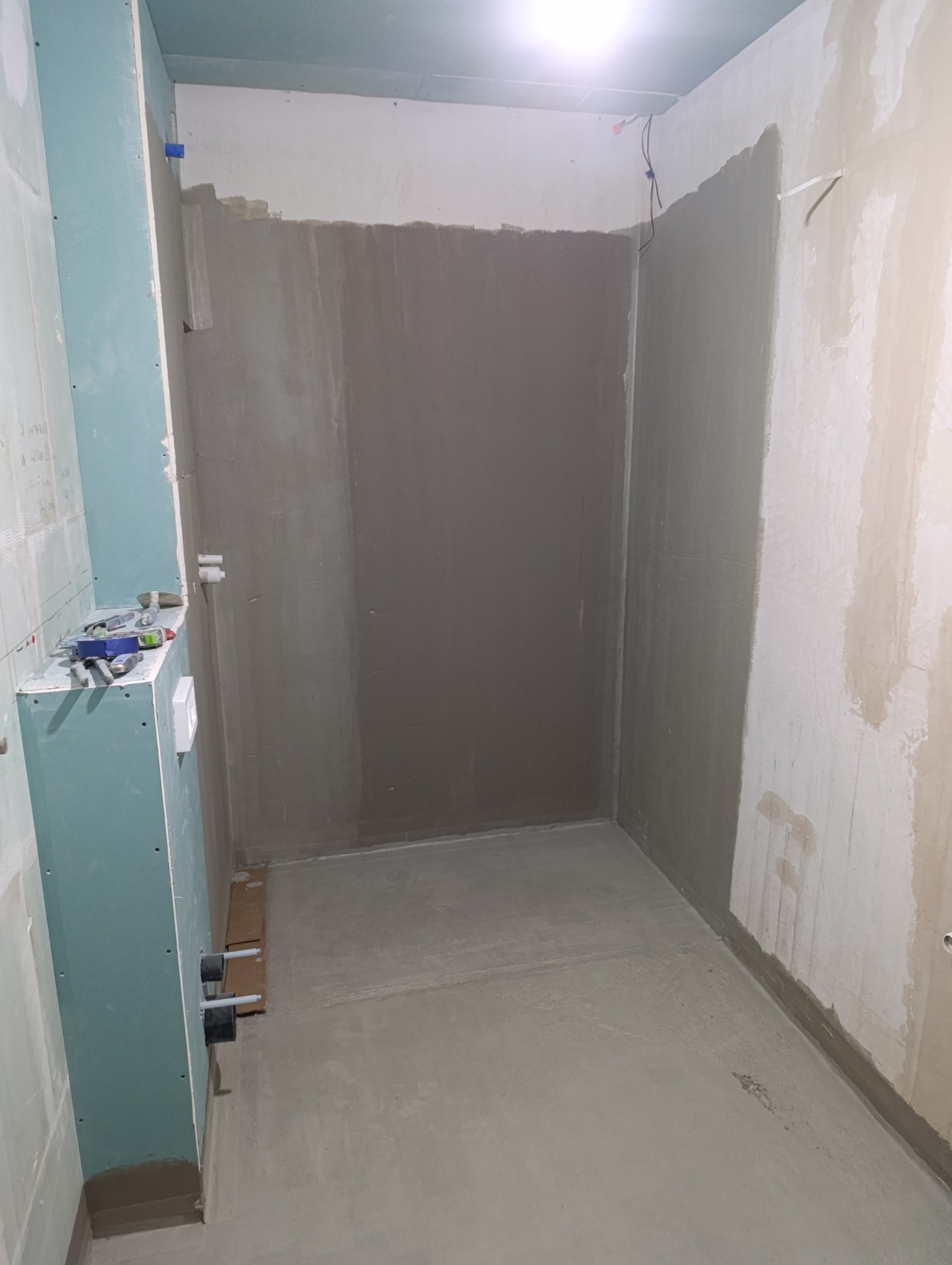 Wnętrze łazienki w trakcie remontu. Widoczne ściany z nałożoną gładzią, płyta gipsowo-kartonowa z narzędziami i wystające rury. Świeżo wylana wylewka na podłodze.