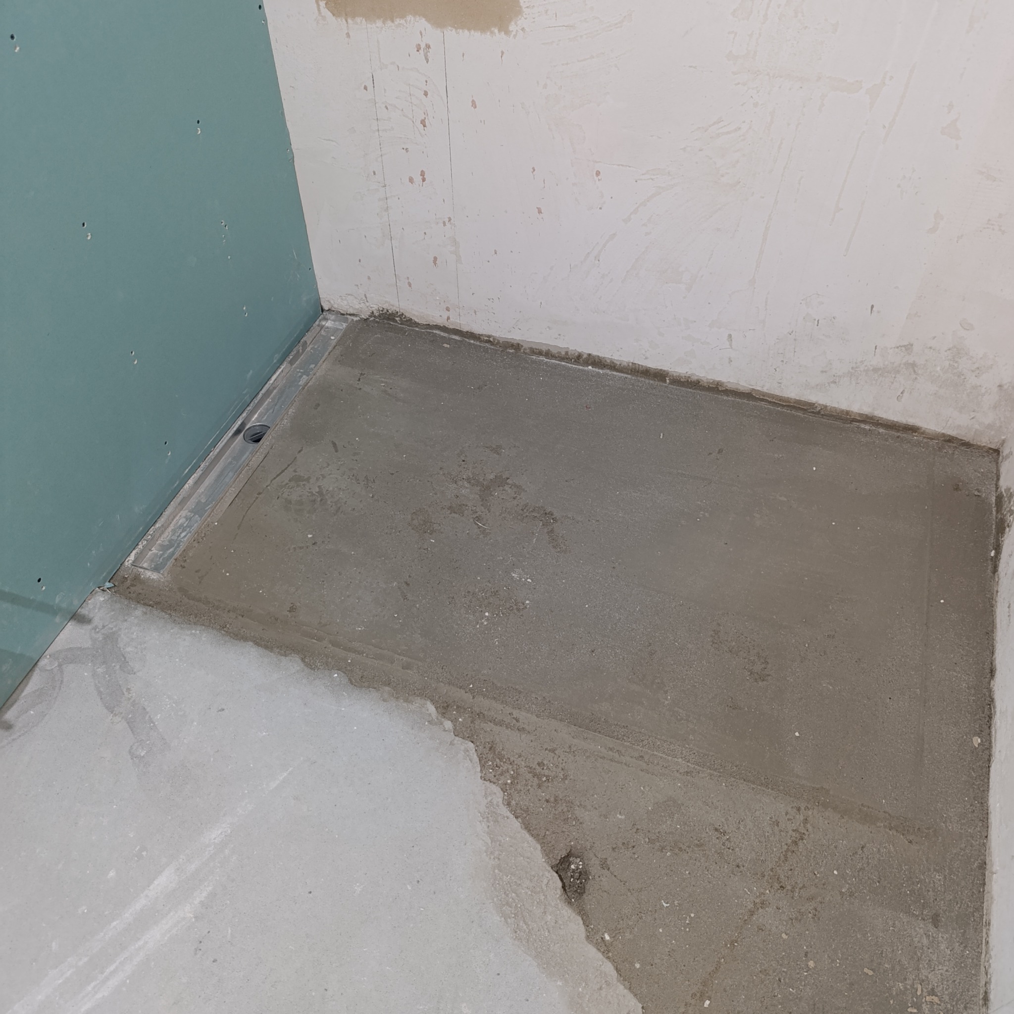 Przygotowana podłoga w łazience z odpływem liniowym, ściany w stanie surowym, płyta GK. Widoczny etap prac przed położeniem płytek. Szary beton i surowe wykończenie.