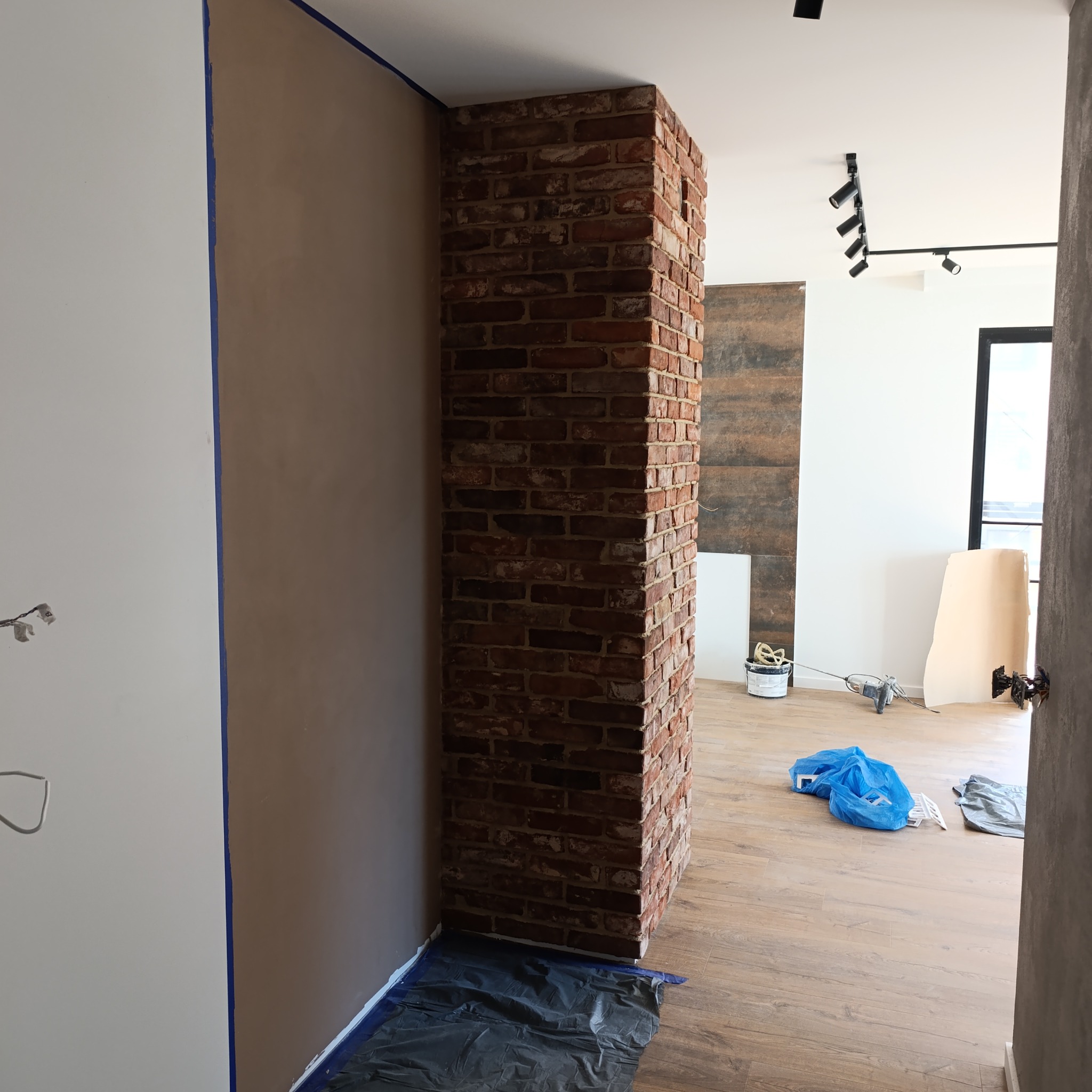 Wykończony filar z cegły klinkierowej w nowym mieszkaniu, zabezpieczony folią malarską. Ściany przygotowane do malowania, widoczne taśmy malarskie.