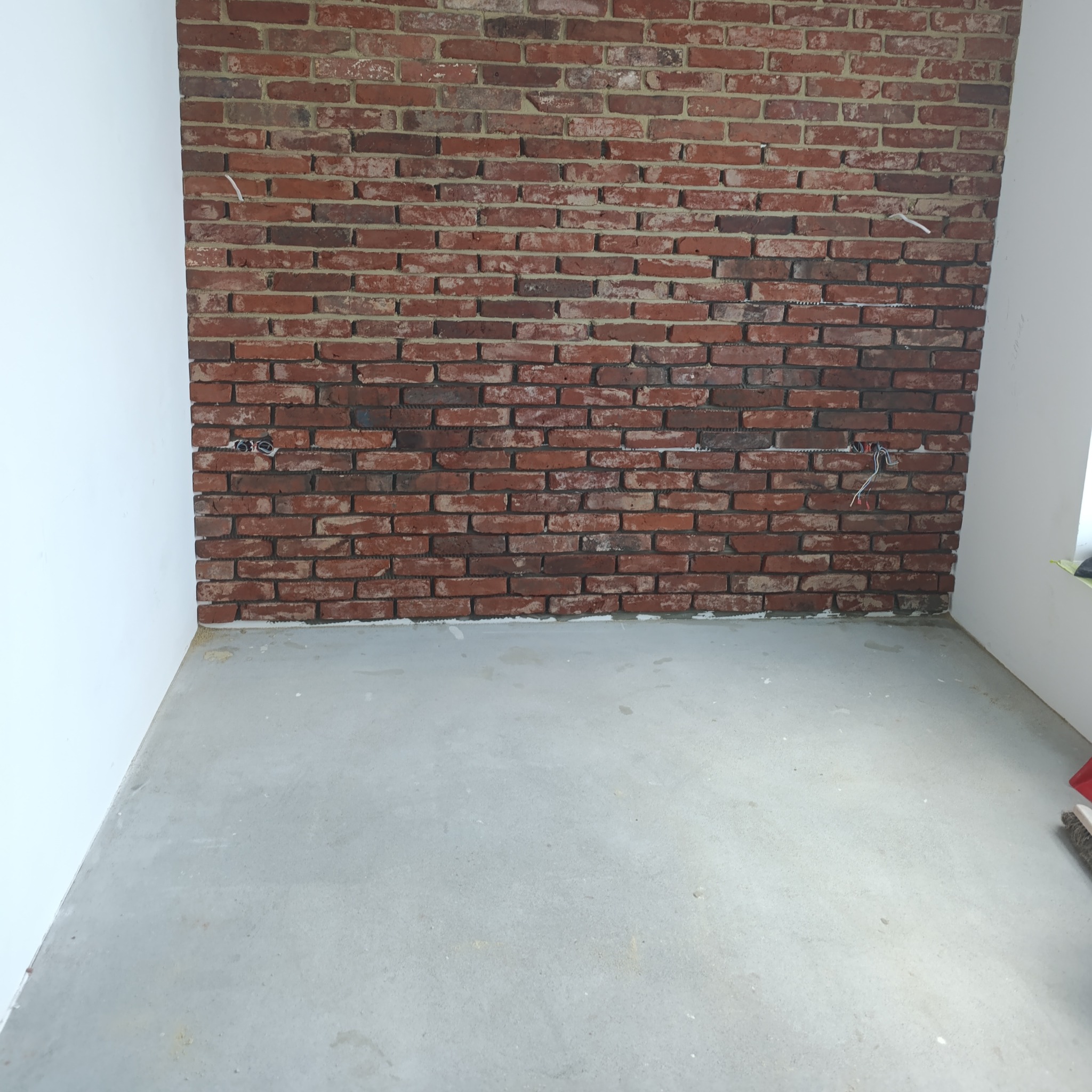 Ściana z czerwonej cegły w surowym wnętrzu, widoczne przewody elektryczne i surowy beton na podłodze, białe ściany po bokach. Nietypowe wykończenie wnętrza.