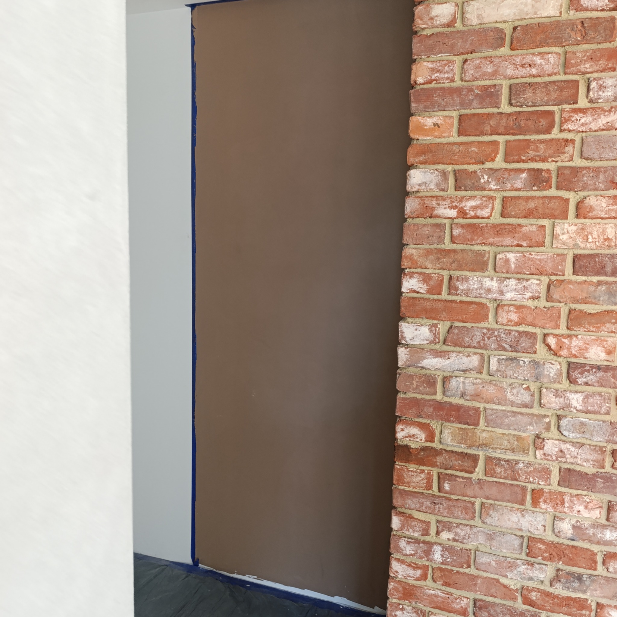 Ściana po nałożeniu gładzi, zabezpieczona taśmą malarską, sąsiadująca z czerwoną ścianą z cegły. Widoczny fragment białej, wykończonej ściany w tle.