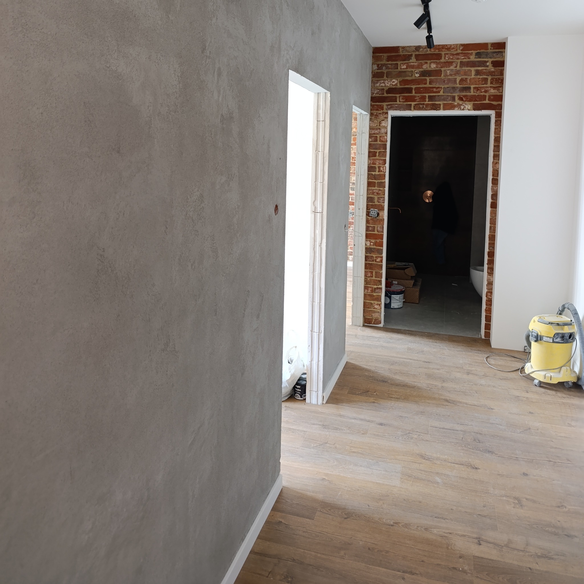 Szara ściana z efektem dekoracyjnym, drewnopodobne panele podłogowe, fragment ściany z cegły i odkurzacz w tle. Widok na wykończone wnętrze.