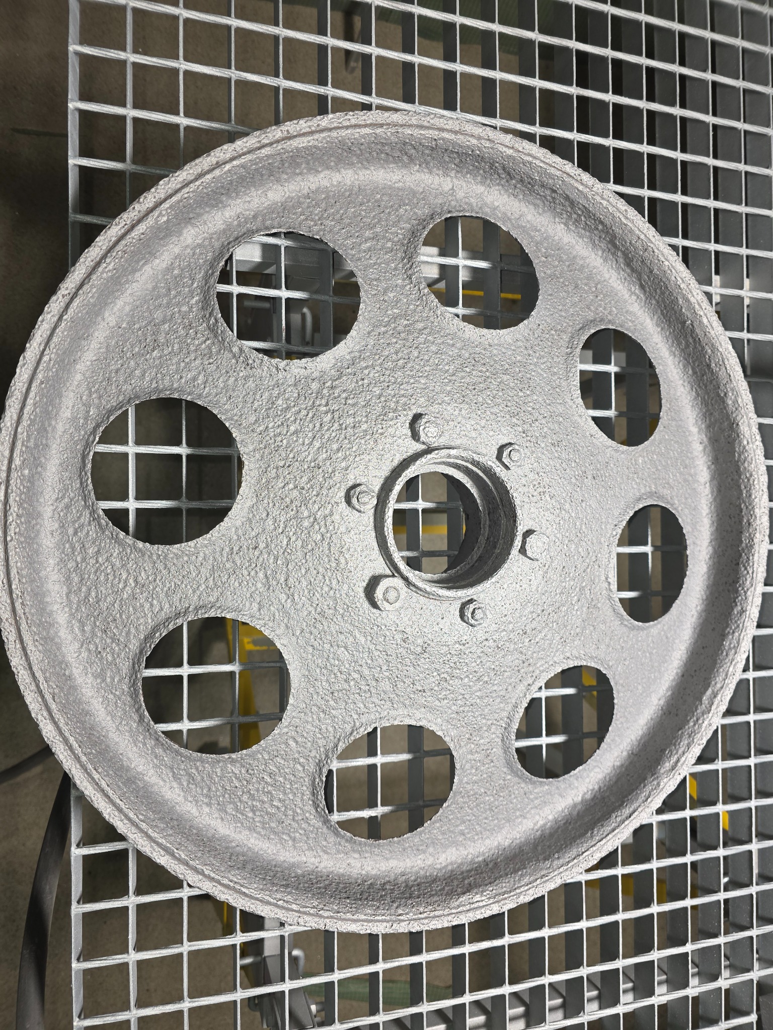 Oczyszczone ścierniwem, metalowe koło z otworami, leżące na metalowej kratownicy. Szorstka, srebrzysta powierzchnia po obróbce.