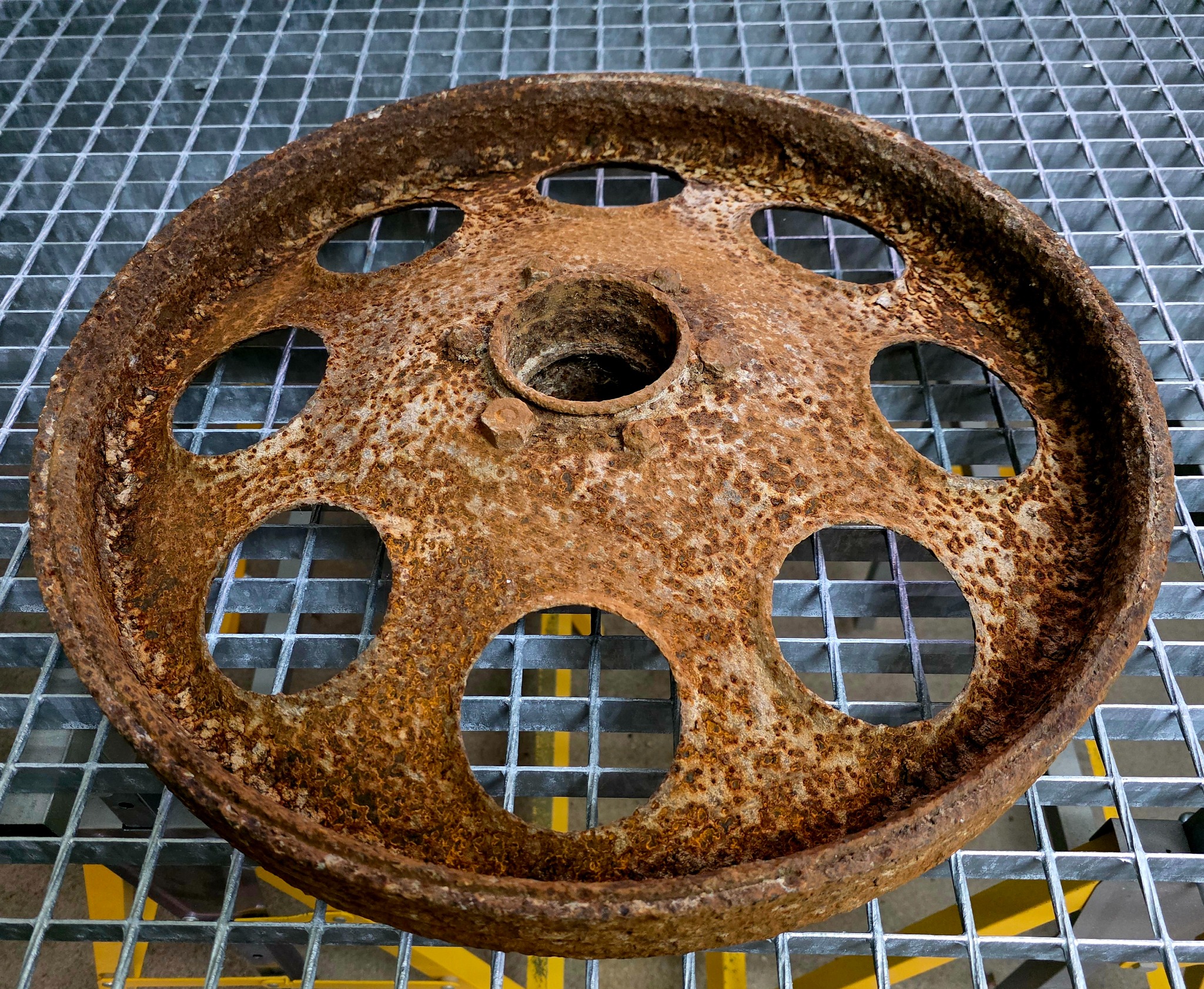 Zardzewiałe, okrągłe koło z otworami, leżące na metalowej kracie. Widoczne ślady korozji i nierówności powierzchni. Detal z bliska, ukazujący strukturę rdzy.