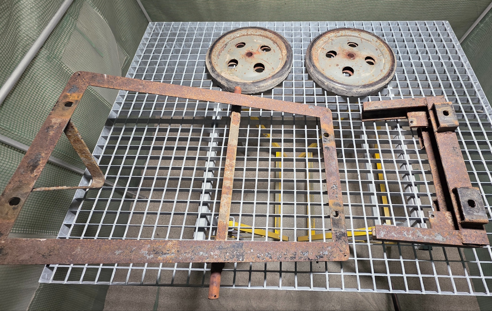 Zardzewiałe, metalowe elementy (rama, koła, wspornik) na metalowej kracie. Widoczne ślady korozji i przygotowania do obróbki. Detal z bliska.