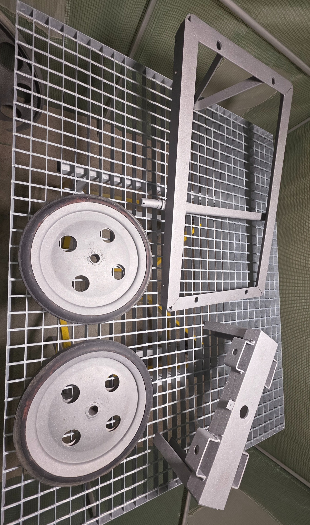 Elementy metalowe (koła, rama, wspornik) po piaskowaniu, leżące na metalowej kratownicy. Widoczne ślady po procesie czyszczenia.