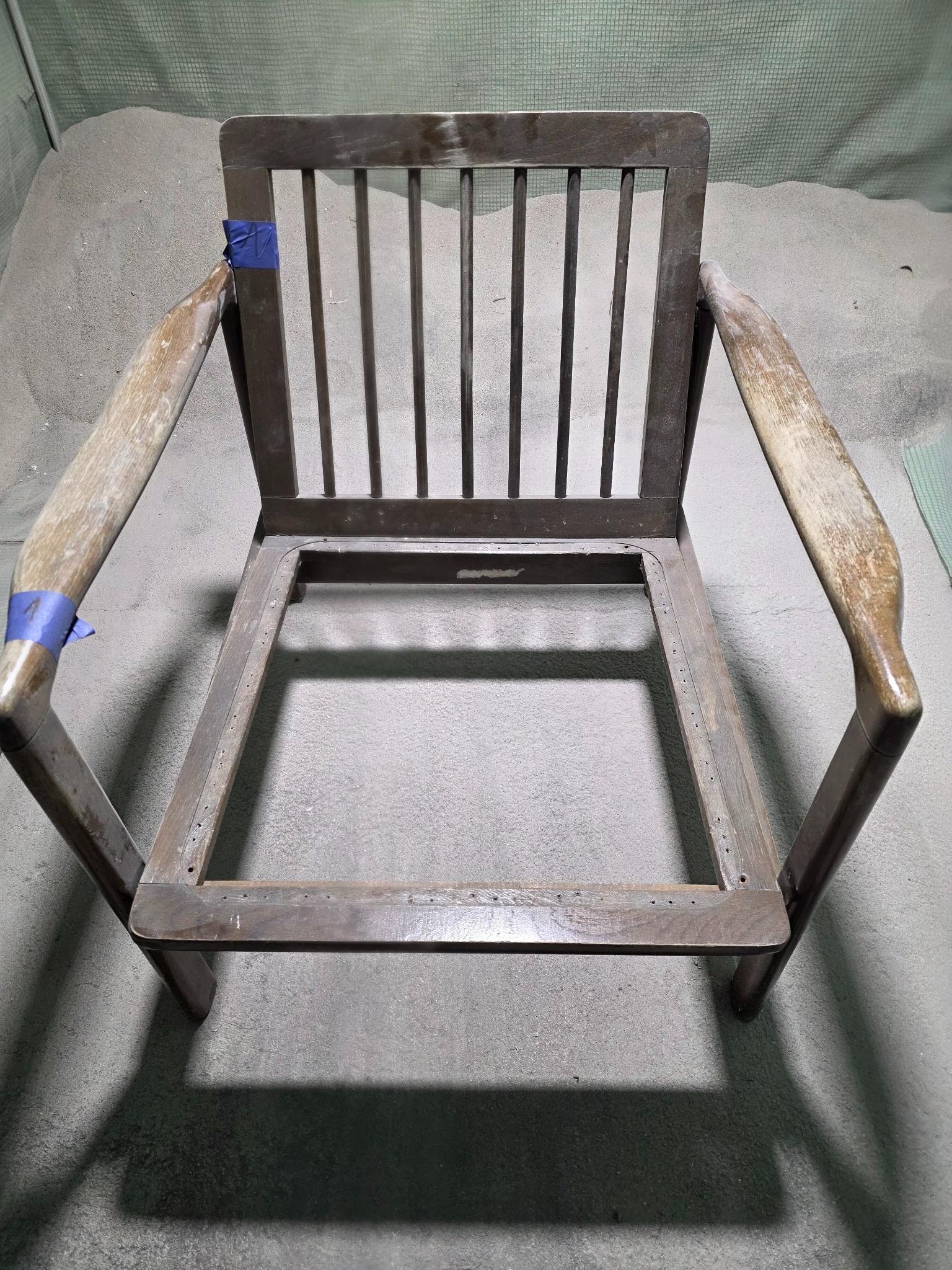 Drewniany szkielet krzesła przed renowacją, z fragmentami taśmy maskującej, na tle szarego podłoża. Widoczne ślady użytkowania i przygotowania do obróbki.